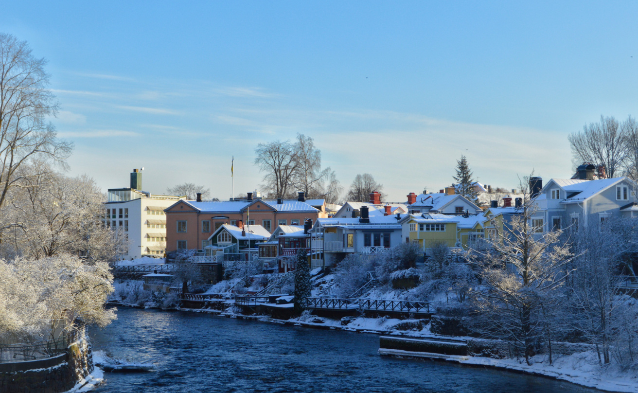 Vintervy över en å i Gävle med snötäckta träd och färgglada trähus längs strandkanten. Himlen är klar och blå, och solen lyser över byggnaderna och det strömmande vattnet.