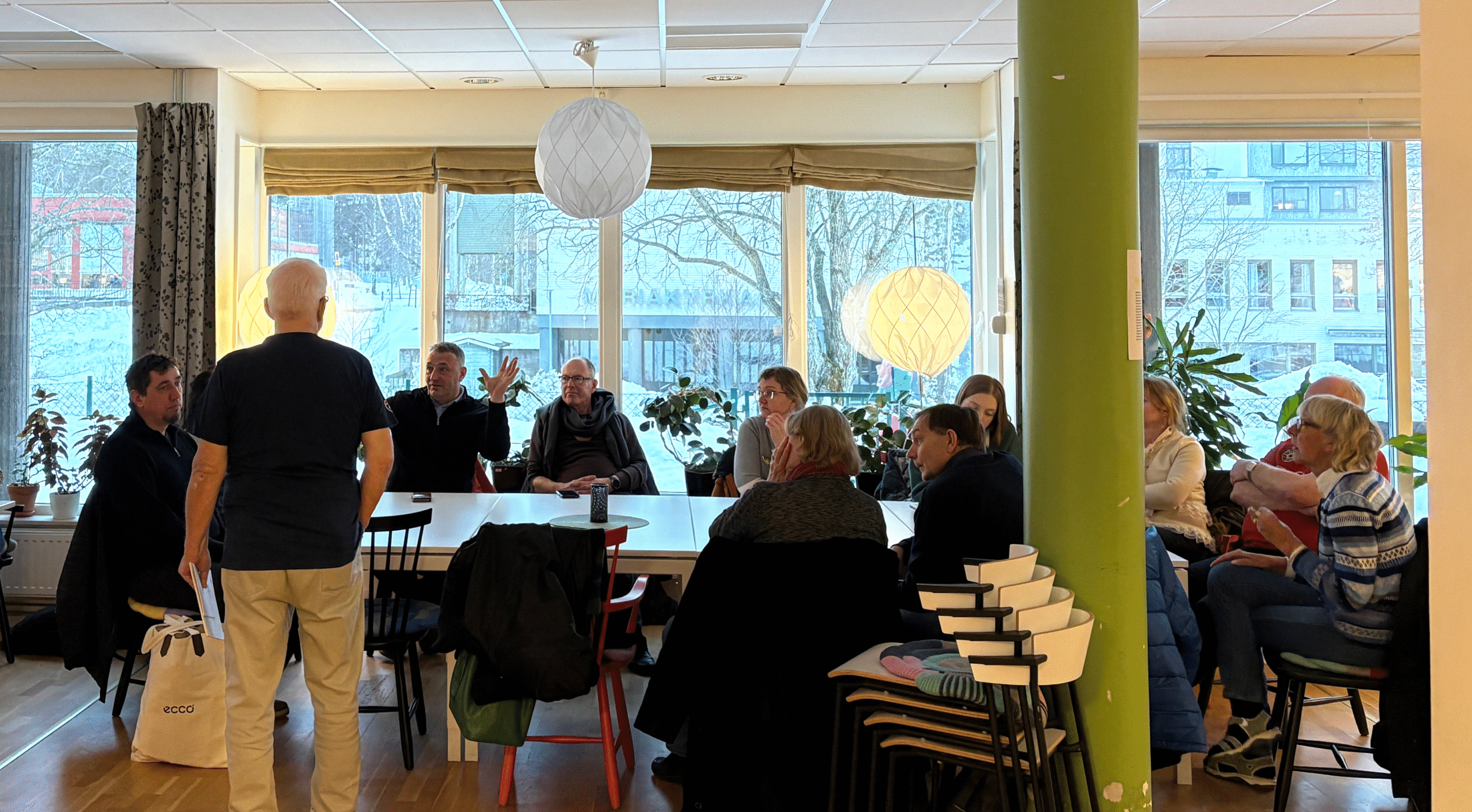 En grupp personer sitter samlade runt ett bord i ett ljust gemensamhetsrum med stora fönster och utsikt över en vintermiljö. En äldre man står upp och talar medan delegationen lyssnar.
