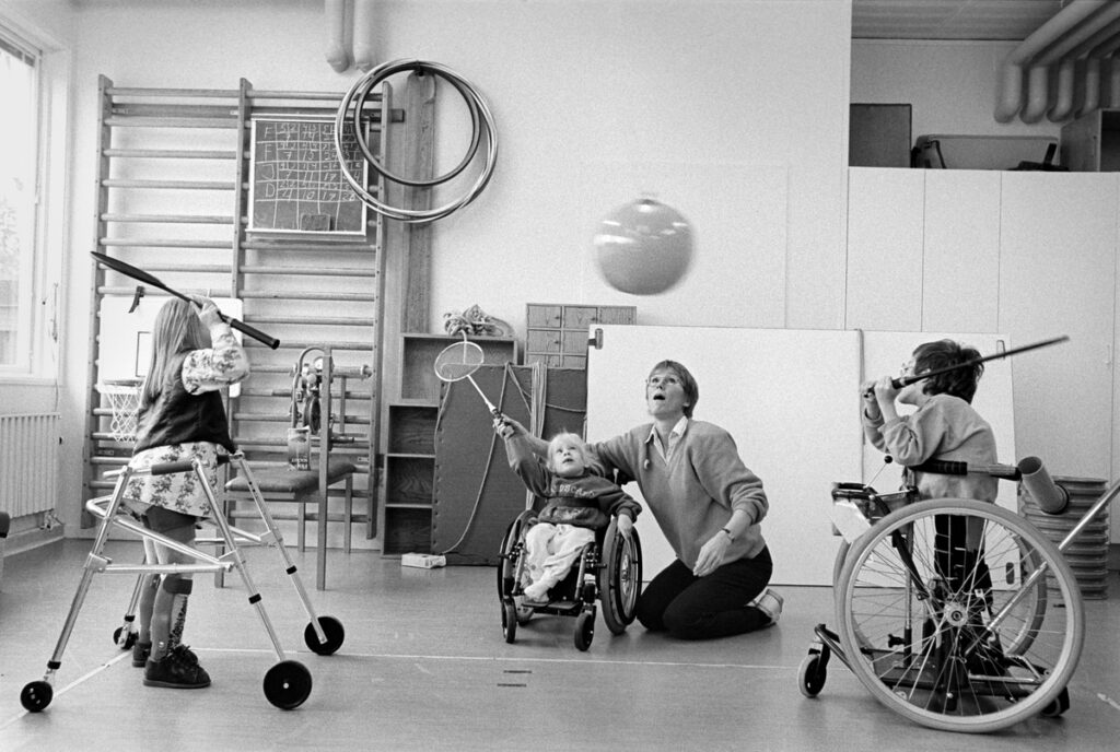 Lärare och tre barn slår på flygande ballong med badmintonracketar. Ett barn sitter i rullstol och två står med hjälpmedel. Fotot är svartvitt.