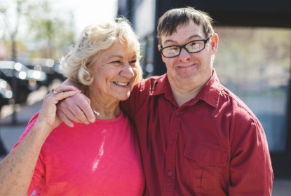 An older woman and a man are standing close together outdoors, smiling at the camera. The woman is wearing a bright pink top, and the man is wearing glasses and a red shirt. They have their arms around each other, and sunlight creates a warm atmosphere in the background.