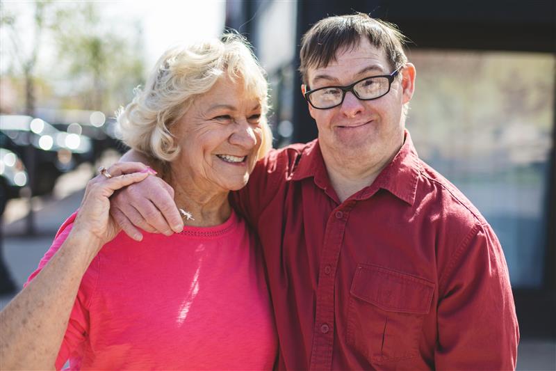 An older woman and a man are standing close together outdoors, smiling at the camera. The woman is wearing a bright pink top, and the man is wearing glasses and a red shirt. They have their arms around each other, and sunlight creates a warm atmosphere in the background.