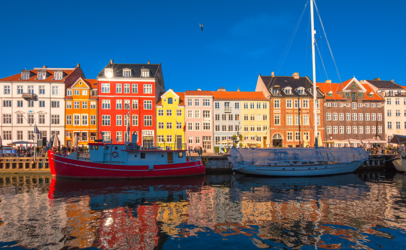 Colourful historic buildings and boats along the waterfront in Nyhavn, Copenhagen, reflected in the water under a clear blue sky.