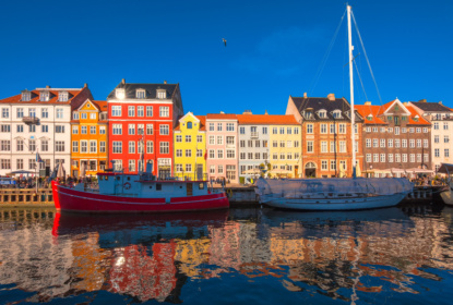 Colourful historic buildings and boats along the waterfront in Nyhavn, Copenhagen, reflected in the water under a clear blue sky.