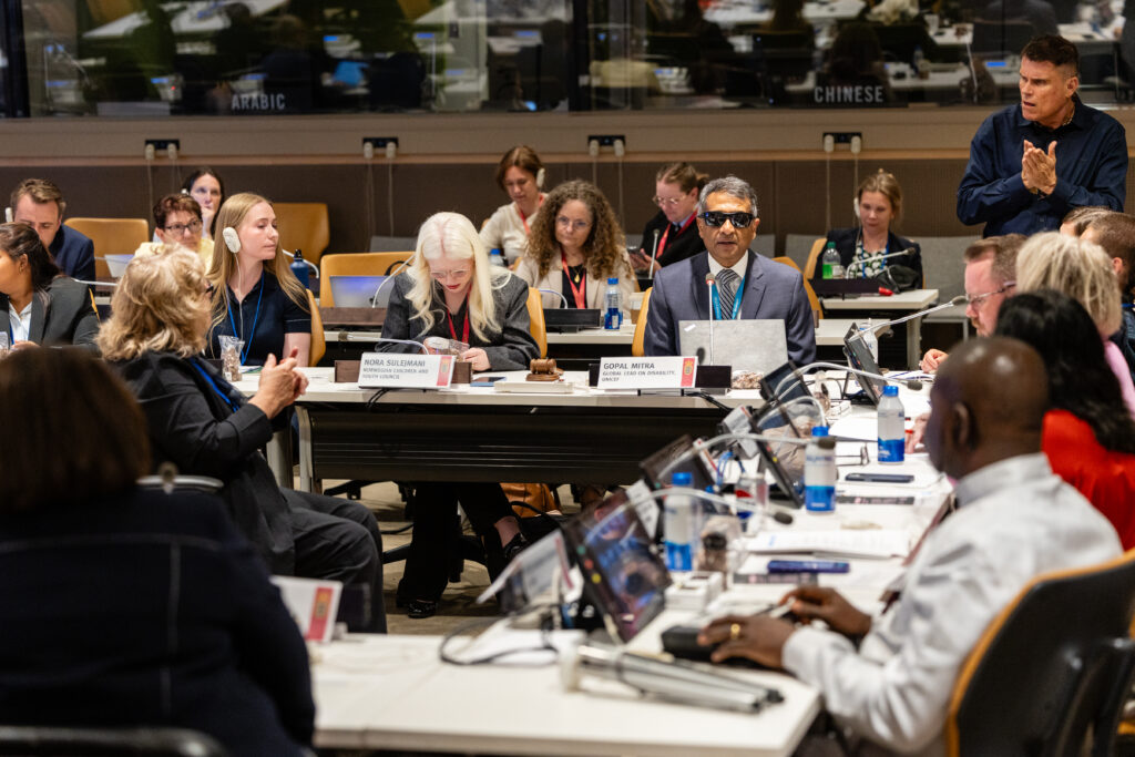 A conference room in the UN Headquarters, Gopal Mitra giving a speech at a conference table. Other participants and audience listening.