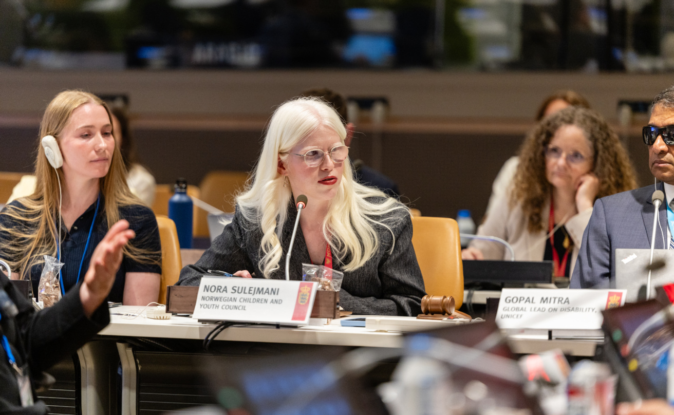 Nora Sulejmani sitting at a conference table and giving a speech. Audience listening behind her. Gopal Mitra and Ninna Simonsen sitting on opposite sides of Nora.