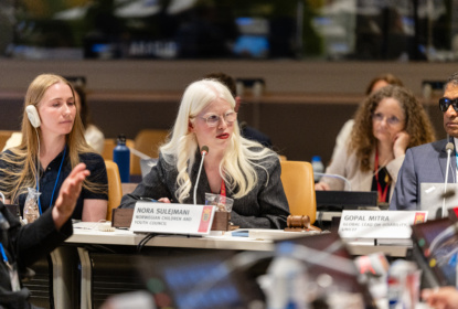 Nora Sulejmani sitting at a conference table and giving a speech. Audience listening behind her. Gopal Mitra and Ninna Simonsen sitting on opposite sides of Nora.