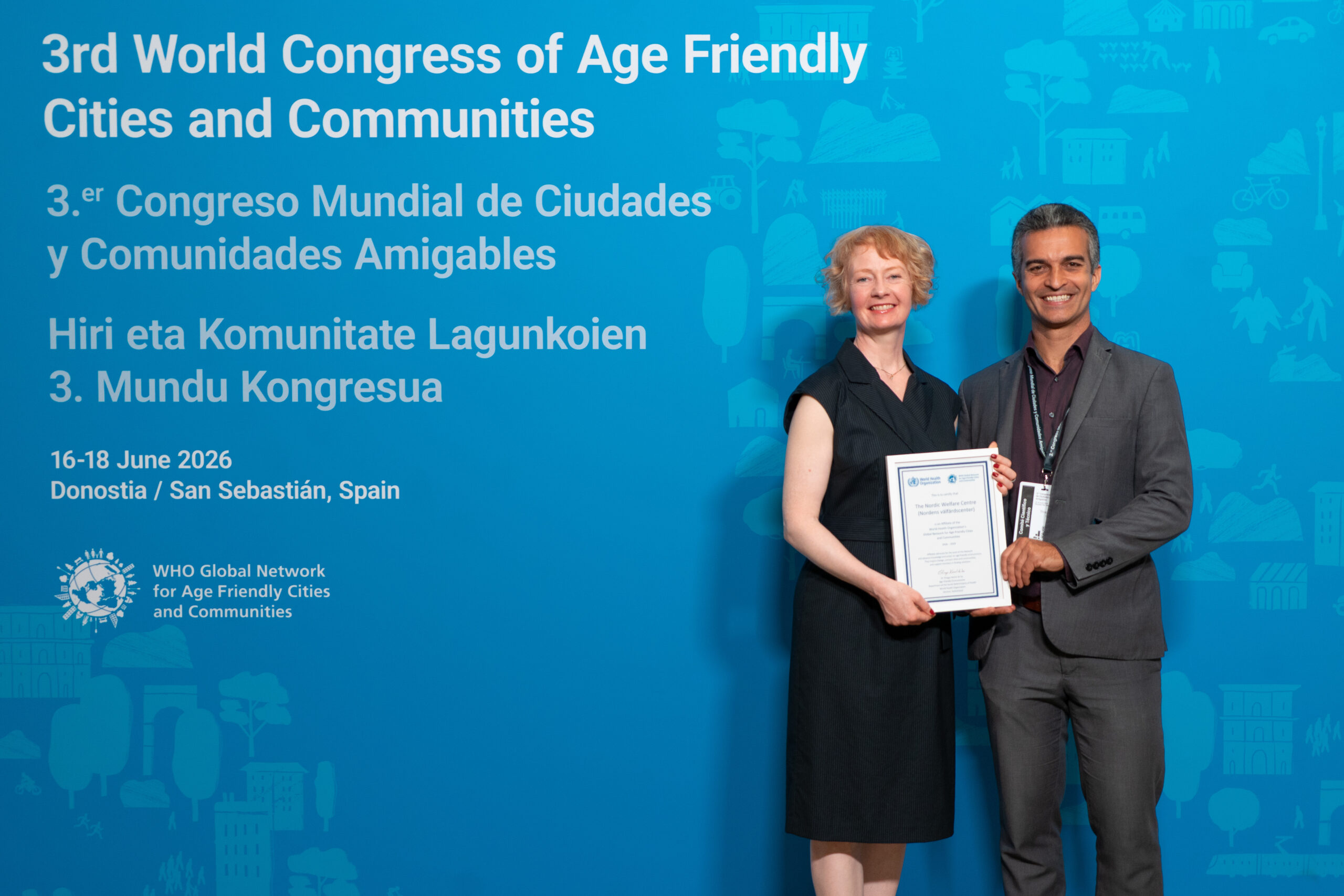 Two conference participants stand in front of a blue backdrop at the 3rd World Congress of Age-friendly Cities and Communities in San Sebastián, Spain, holding a certificate recognizing the Nordic Welfare Centre as an affiliate of the WHO Global Network for Age-friendly Cities and Communities. The backdrop displays the congress title, dates (16–18 June 2026), location, and the WHO Global Network logo.