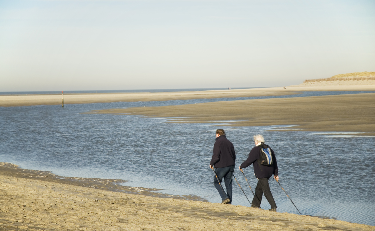 Two older adults walking with Nordic walking poles along a wide sandy beach next to a calm tidal channel. Sand dunes and the open sea stretch into the distance beneath a clear sky.