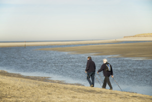 Two older adults walking with Nordic walking poles along a wide sandy beach next to a calm tidal channel. Sand dunes and the open sea stretch into the distance beneath a clear sky.