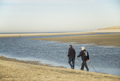 Two older adults walking with Nordic walking poles along a wide sandy beach next to a calm tidal channel. Sand dunes and the open sea stretch into the distance beneath a clear sky.