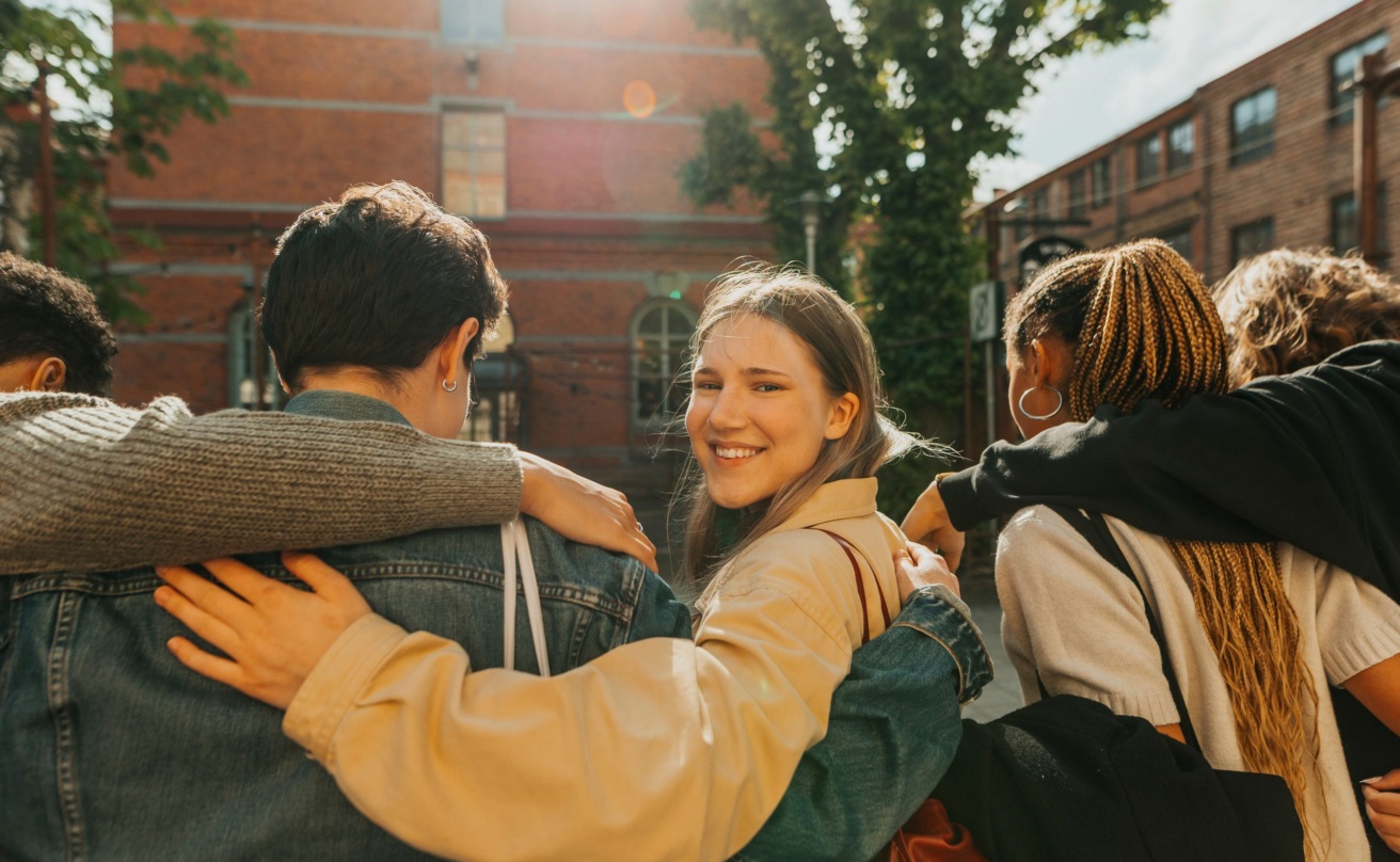 portrait-of-smiling-young-woman-looking-over-shoulder-while-walking-with-arm-around-friends-at-street