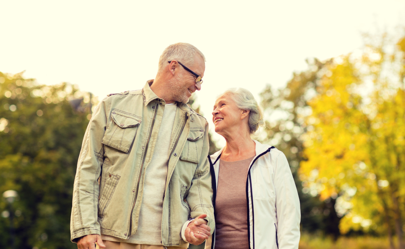 An older couple walks hand in hand through a park, smiling warmly at each other. Trees with autumn foliage are visible in the background.