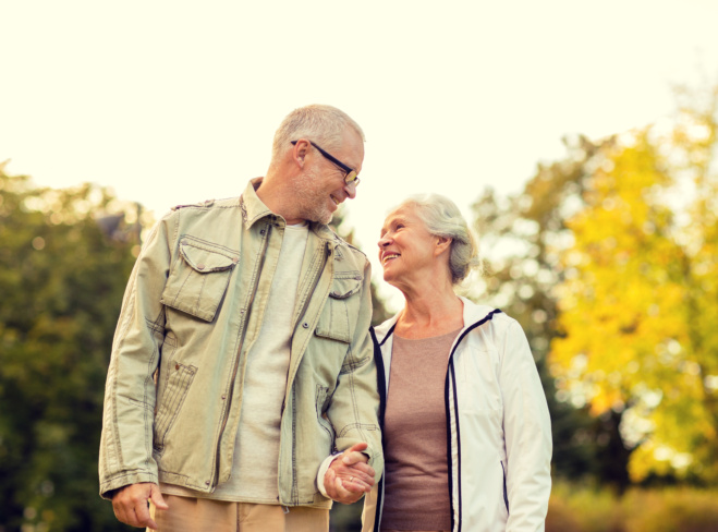 An older couple walks hand in hand through a park, smiling warmly at each other. Trees with autumn foliage are visible in the background.