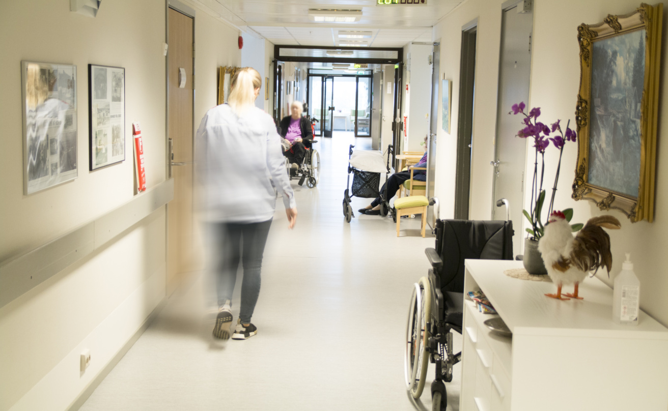 Korridor på ett äldreboende där en person går genom korridoren. Äldre personer sitter längre bort, några med rullstol och rollator.