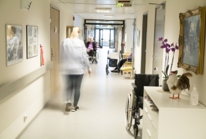 Korridor på ett äldreboende där en person går genom korridoren. Äldre personer sitter längre bort, några med rullstol och rollator.
