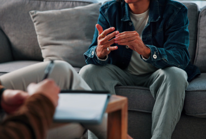 A person sits on a grey sofa, gesturing with their hands during a conversation. Another person sits in the foreground with a tablet on their lap.